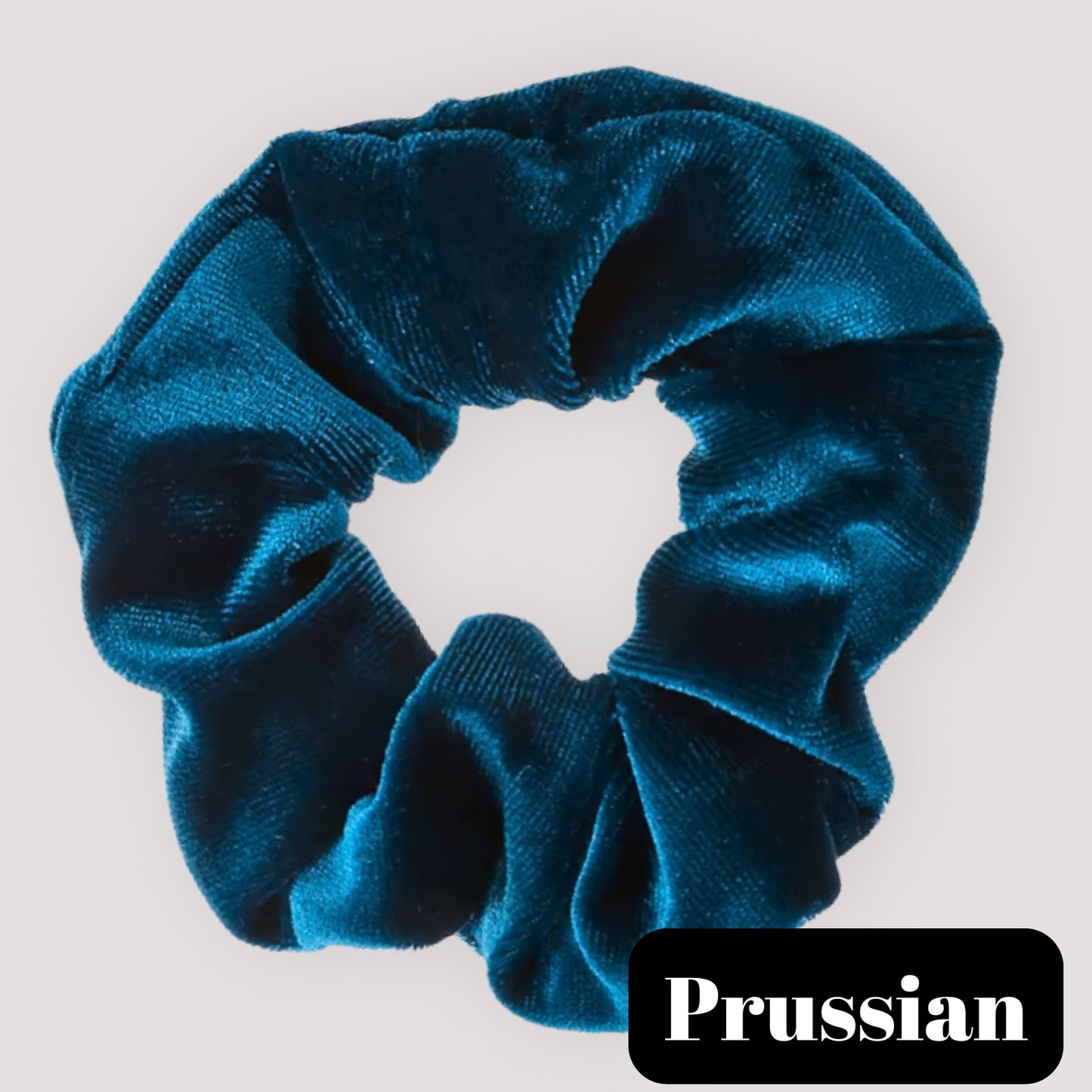 A single Prussian dark teal blue handmade velvet scrunchie for damage-free hair styling on a light background, featuring a black label reading 'Prussian'.