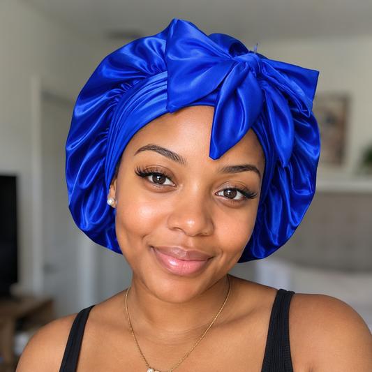 A woman wearing a shiny royal blue satin turban headwrap with a large bow, shown in a close-up portrait against a softly blurred indoor background.