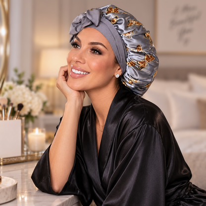 A woman wearing a gray satin bonnet with gold butterfly patterns and a black satin robe, posing in a bright, modern bedroom setting.