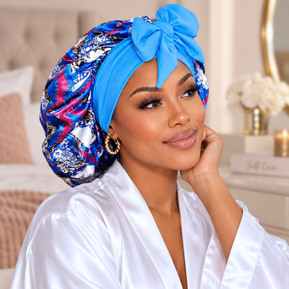 A woman wearing a bright blue satin bonnet with a large bow and a matching patterned bonnet underneath, paired with a white satin robe in a softly lit bedroom setting.