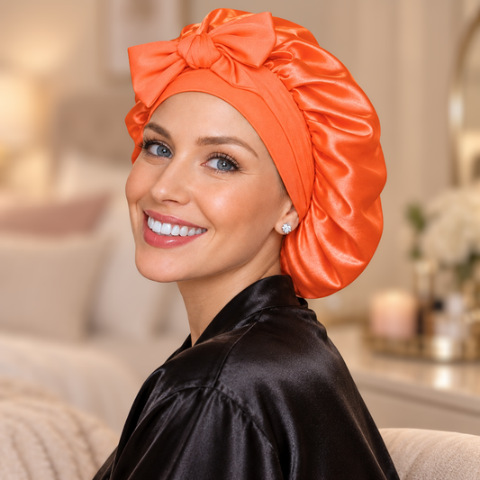 A smiling woman wearing a shiny bright orange satin turban-style headwrap with a large bow, paired with a black satin robe, photographed in a softly lit indoor setting.