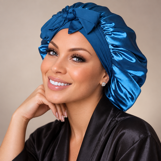 A smiling woman wearing a shiny peacock blue satin turban bonnet and a black satin robe, photographed against a soft beige studio background.