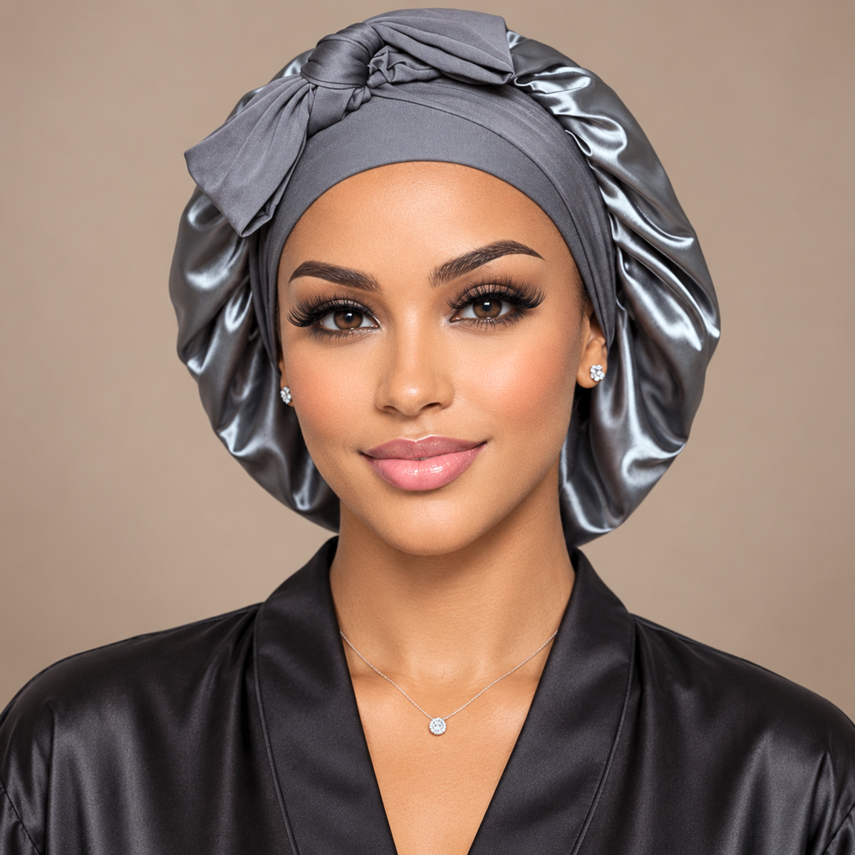 A woman wearing a shiny dark gray satin turban headwrap and a matching black satin robe, shown from the chest up against a plain beige studio background.