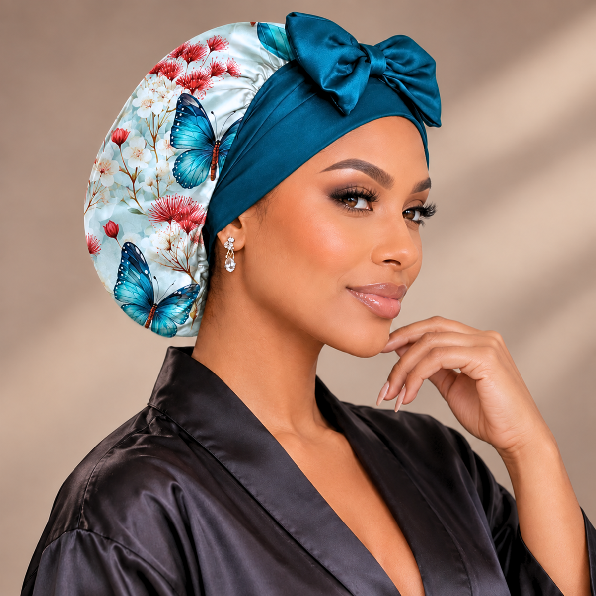 A woman wearing a satin sleep cap with a large bow, featuring a light blue floral and butterfly print on one side and a solid teal blue bow on top. She is wearing a black satin robe against a warm beige background.