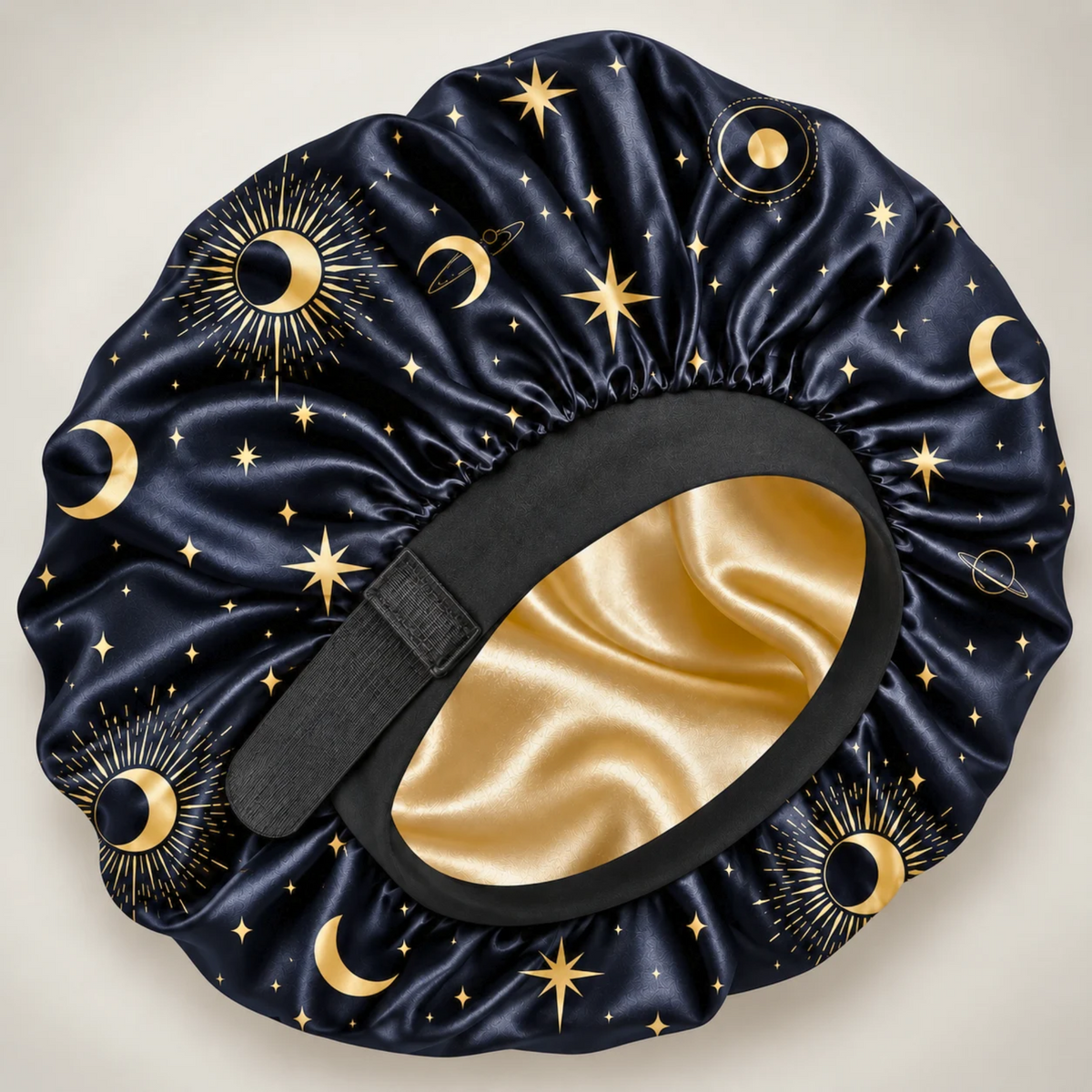 A satin sleep cap or bonnet with a celestial print, featuring a navy blue base with gold stars, crescent moons, and sun motifs, a black elastic band, and a shiny gold interior.